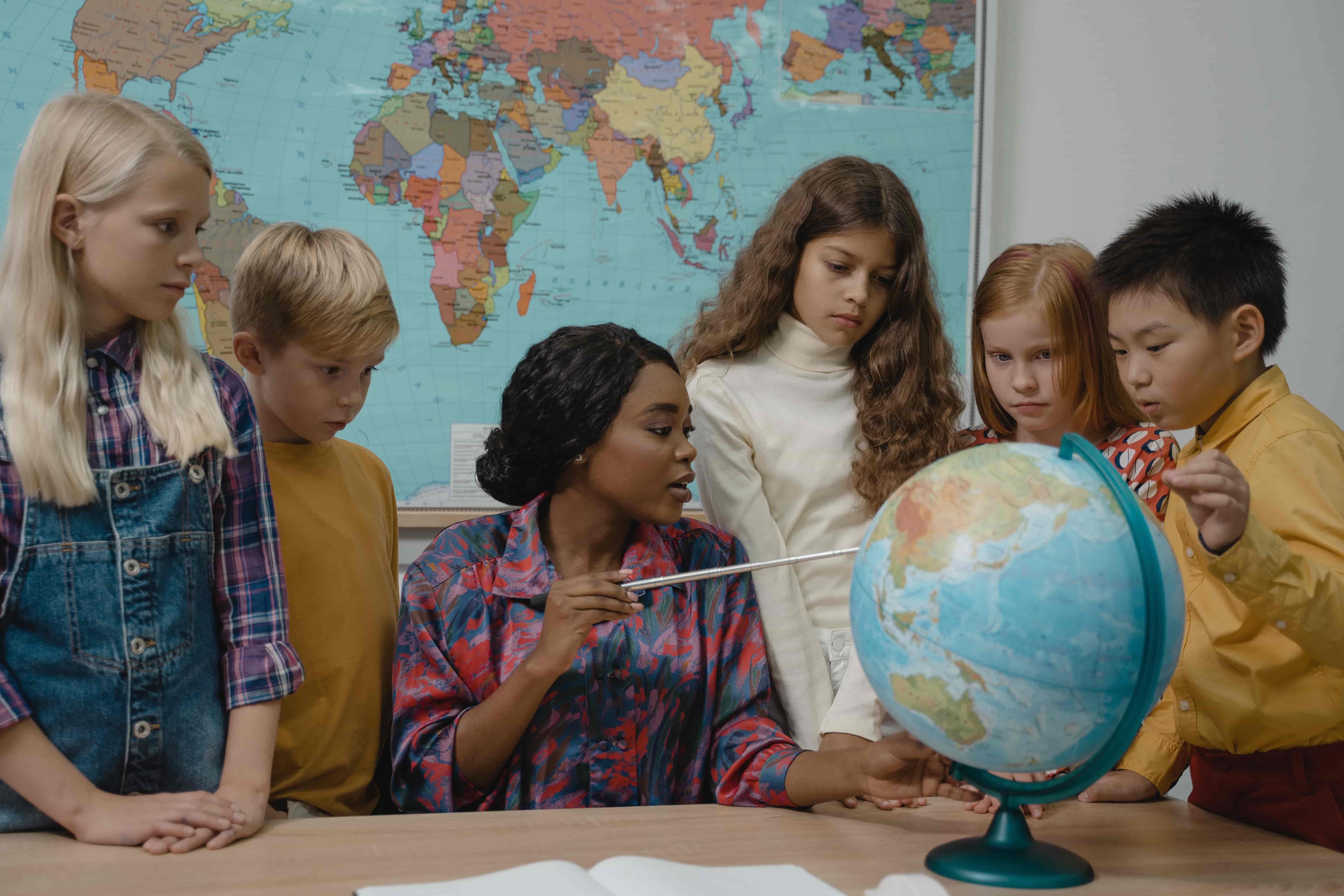 intercultural trainer pointing to globe teaching group of five young, diverse children about different countries and cultures